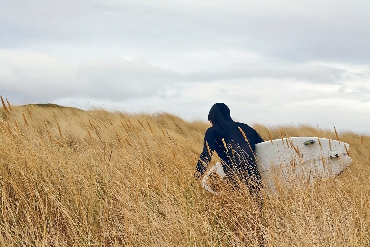 man in wet suit walking through wheat field carrying surfboard