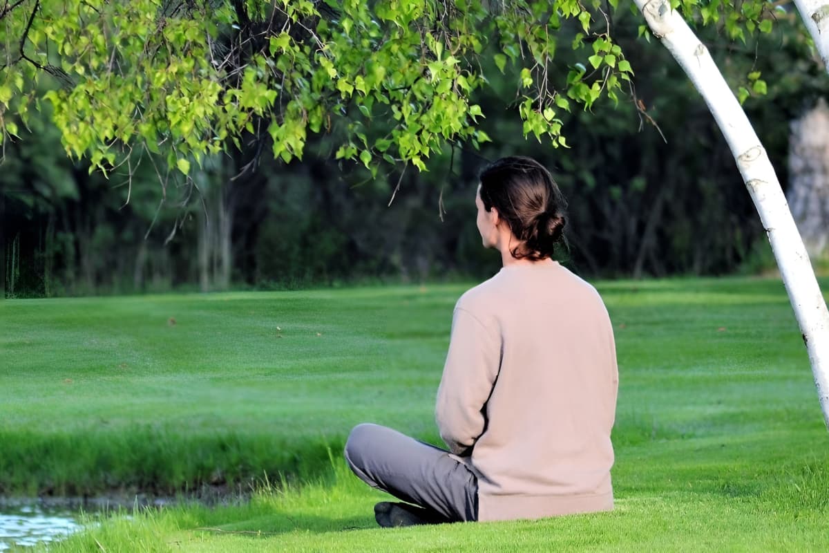 back of woman sitting cross legged by pond and trees